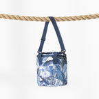 A blue and white tropical leaf print cooler bag with a navy blue shoulder strap, hanging from a thick natural rope against a clean white background.