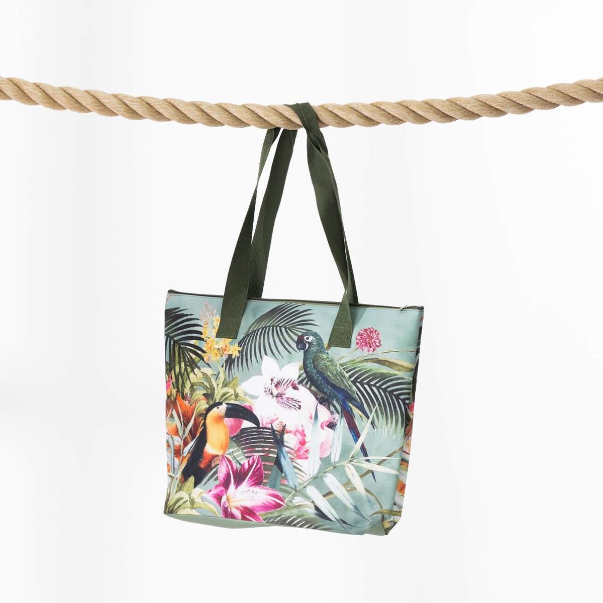 A beach bag with a tropical jungle print featuring toucans, parrots, and palm leaves, hanging from dark green straps over a thick rope against a white background.