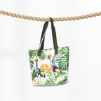 A beach bag with a tropical print of birds, flowers, and leaves, hanging from dark green straps over a thick rope against a white background.