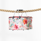 A rectangular folding cooler basket with a vibrant tropical floral print, suspended by a black handle from a thick natural rope against a clean white background.