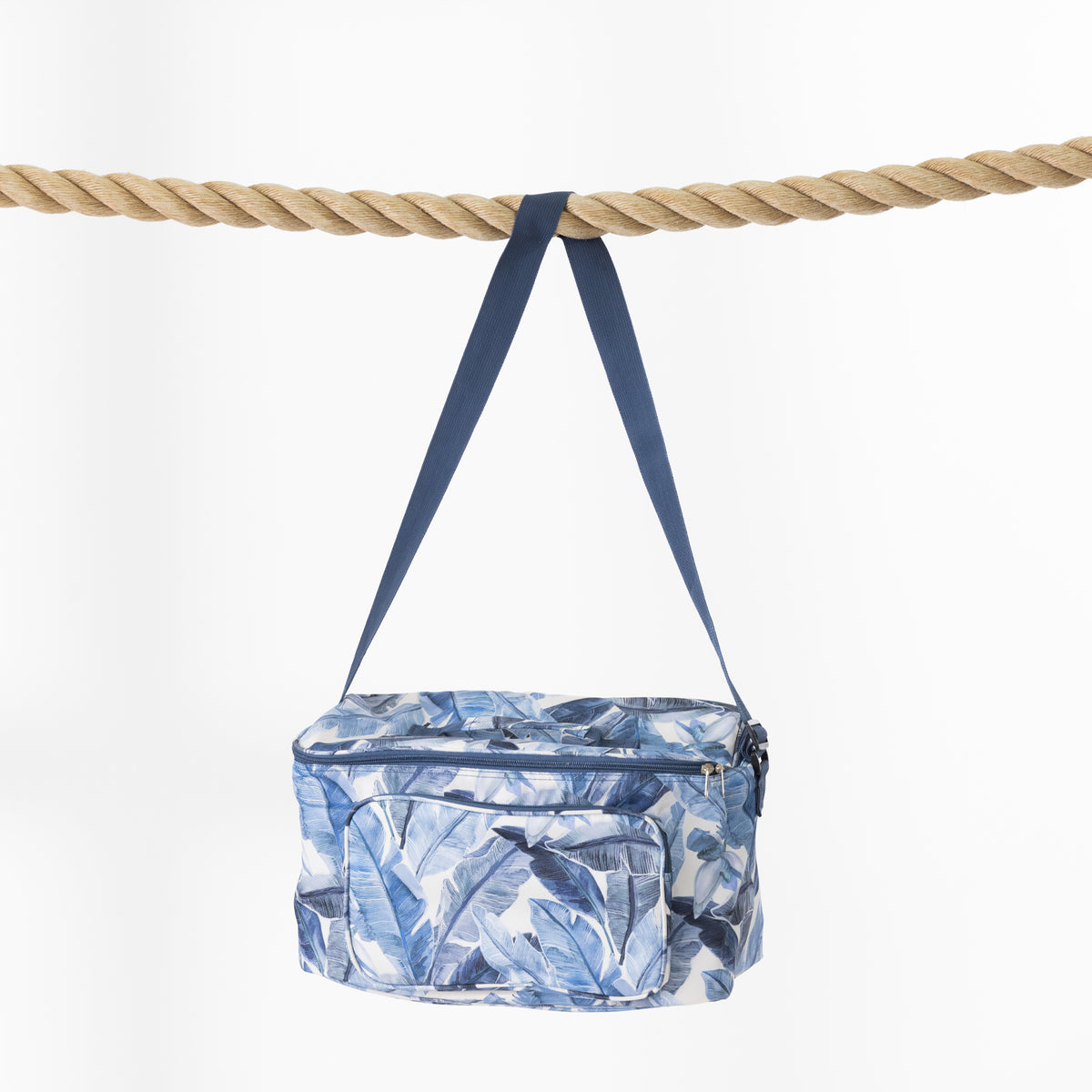 A blue and white tropical leaf print cooler bag with a blue strap, hanging from a thick natural rope against a clean white background.