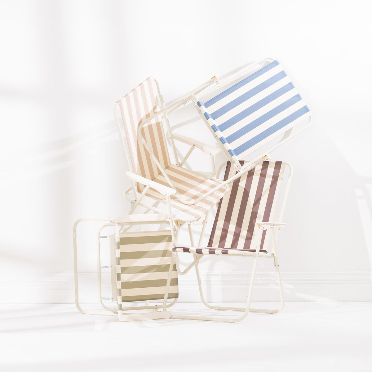 Four striped folding beach chairs in Green Stripe, Blue Stripe, Natural Stripe and Mocha stacked and arranged together on a white studio background.