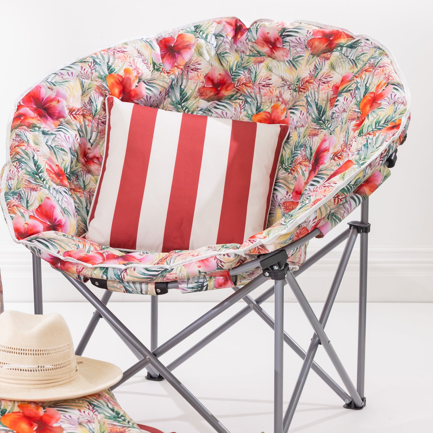 A tropical floral print folding moon chair with a red and white striped throw pillow, set against a clean white background with a straw hat prop in the foreground.