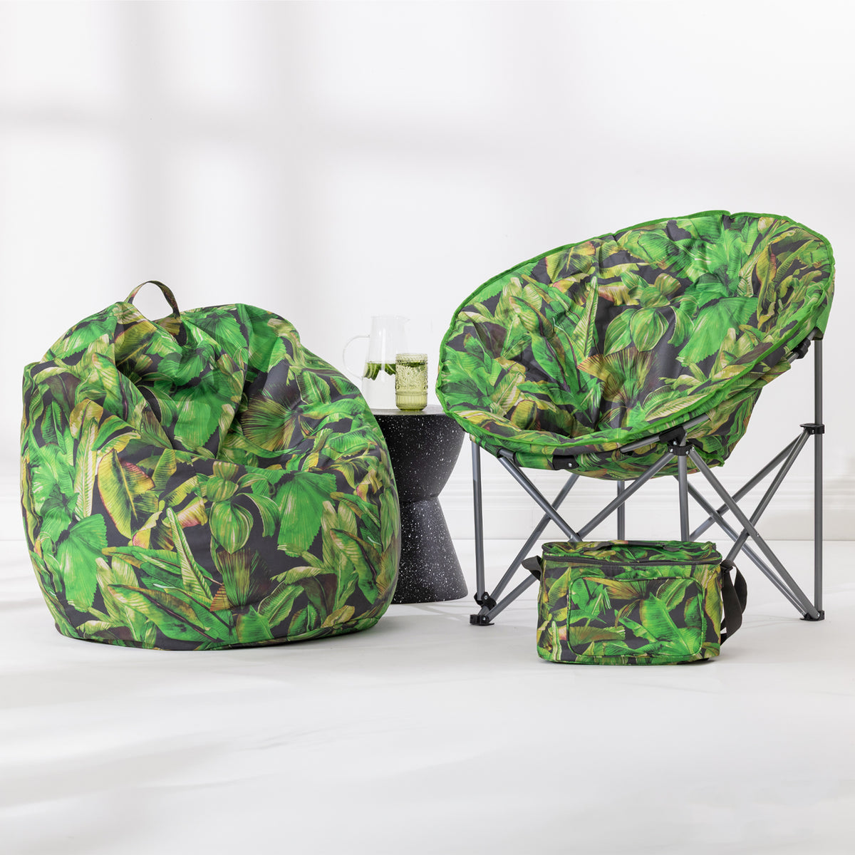 A coordinated outdoor seating set with a green tropical leaf print bean bag chair, a matching folding moon chair, and a small matching cooler bag, displayed on a clean white studio floor with a small dark side table between them.