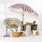 A beach-themed outdoor furniture and accessory set arranged on a white floor against a white wall. The set includes a striped beach umbrella with fringe, a patterned moon chair, a wicker picnic basket, a folding beach trolley with yellow wheels, a striped towel, and sandals.