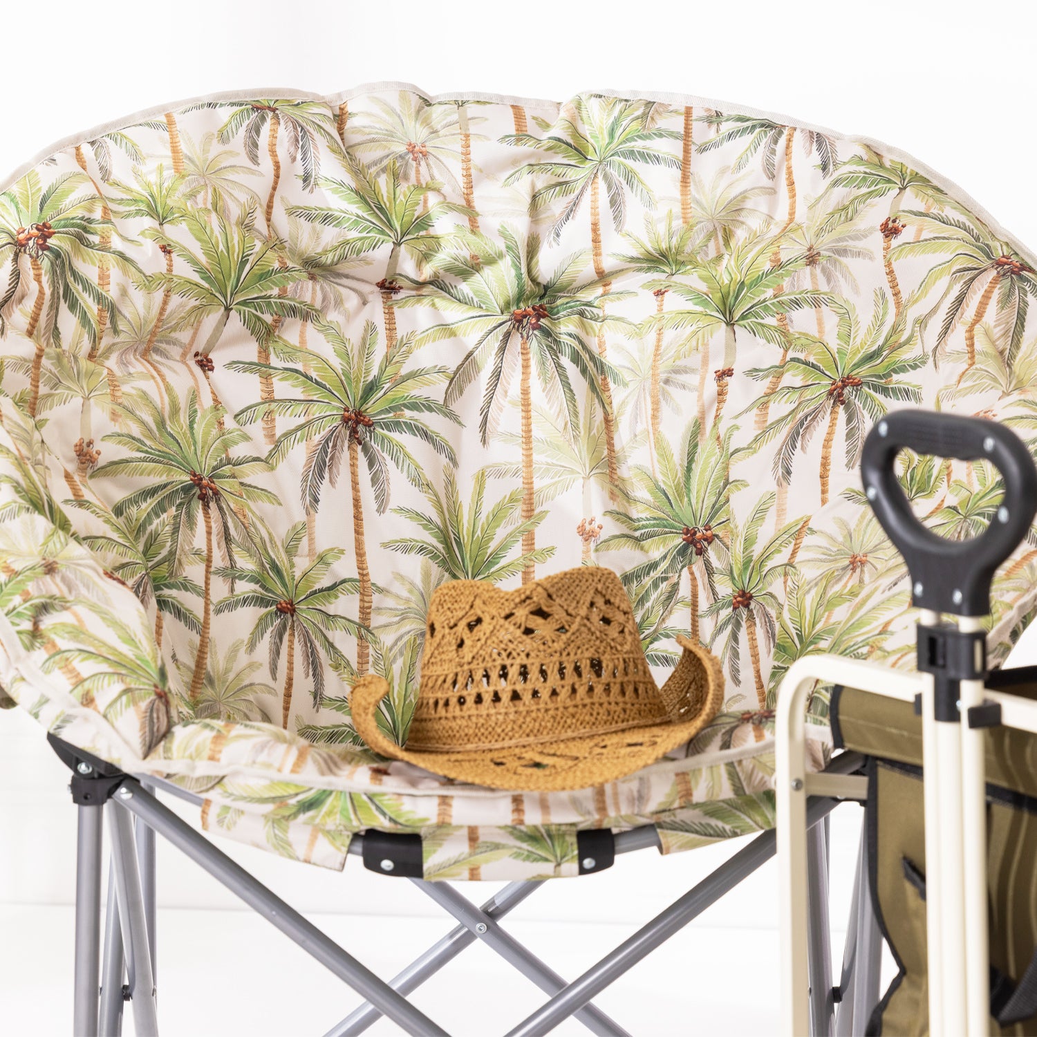 A close up of a round moon chair with a tropical palm tree print cushion, shown with a woven straw hat resting on the seat. 