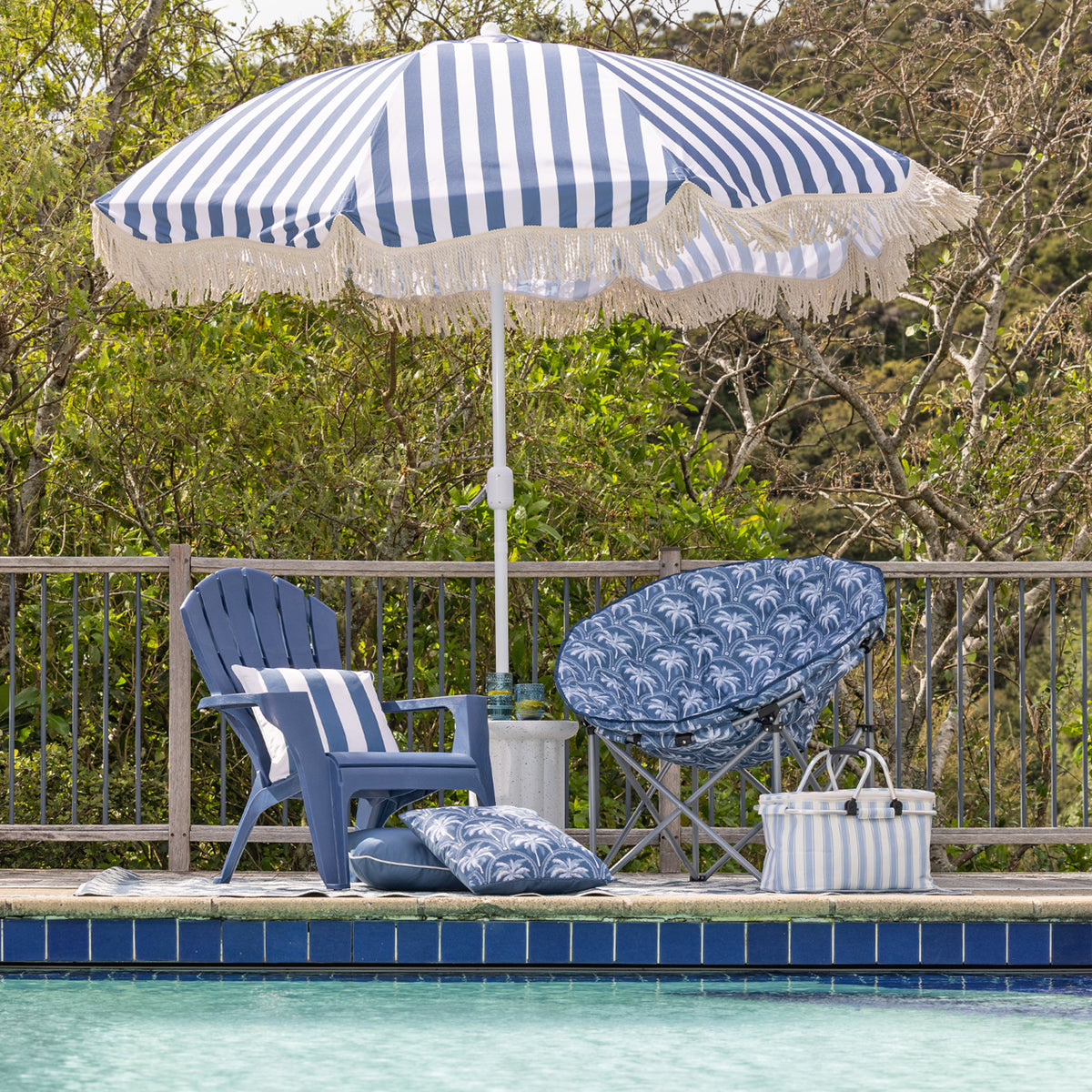 A blue and white striped beach umbrella with fringe stands over a poolside patio setup featuring a blue cape cod chair, a blue patterned lounge chair, matching pillows, and a striped cooler bag.