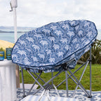 A round outdoor moon lounge chair with a blue and white palm tree patterned cushion, set on a the grass with a scenic ocean view in the background.