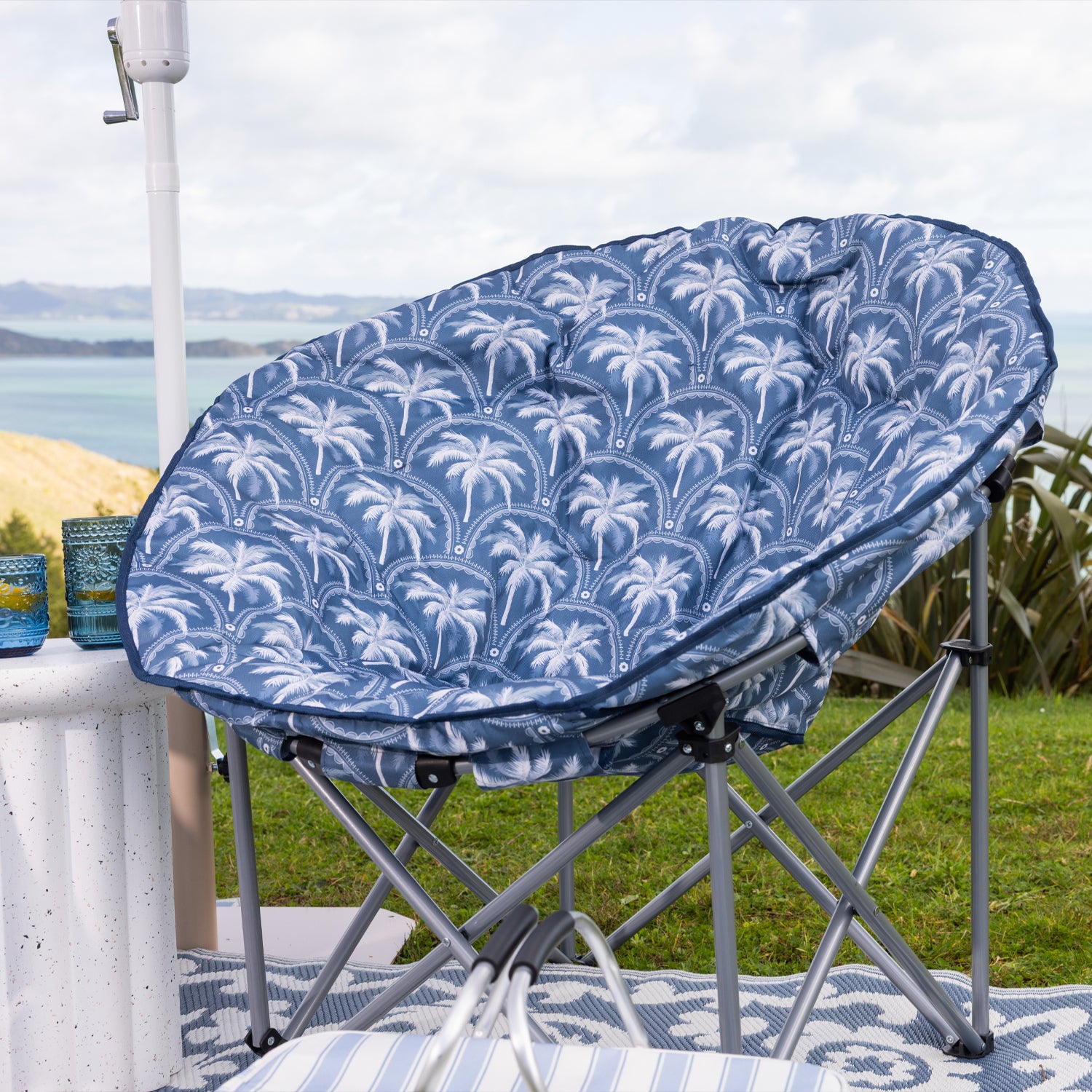 A round outdoor moon lounge chair with a blue and white palm tree patterned cushion, set on a the grass with a scenic ocean view in the background.
