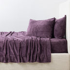 Purple textured sheet set and pillows on a bed against a white wall.