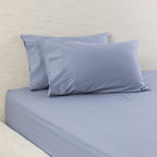 A bed dressed in a smooth, solid blue coloured fitted sheet with two matching pillowcases, set against a textured beige headboard.