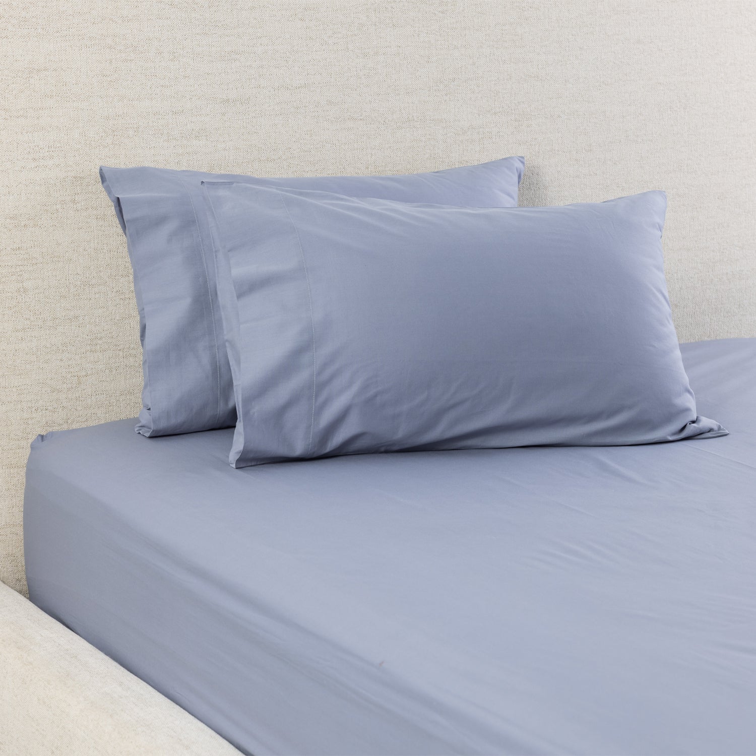 A bed dressed in a smooth, solid blue coloured fitted sheet with two matching pillowcases, set against a textured beige headboard.